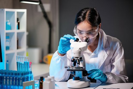 Woman wearing safety glasses, lab coat and blue gloves peering into a microscope