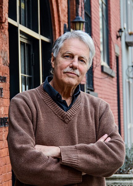 Photo of Desplan, wearing a brown sweater and black collared shirt, standing against a red brick building.