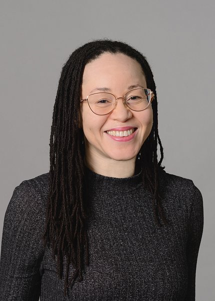 Headshot of West-Mitchell. She is wearing glasses and a black shirt and is standing against a light gray background.