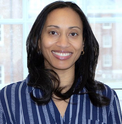 Forde, smiling and wearing a blue and white striped shirt, stands in front of a window. A brick building is visible beyond the window.