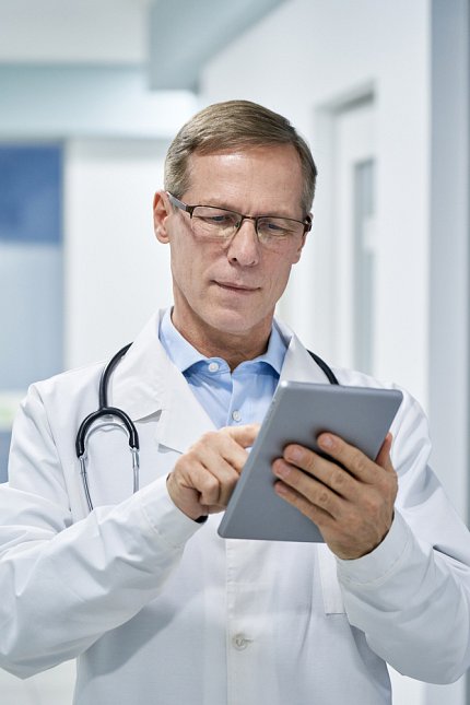 A bespectacled male doctor reads something on a tablet. He wears a white coat and a stethoscope around his neck.