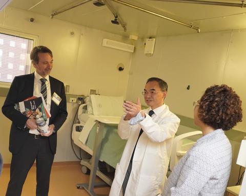 Dr. Chen extends hands as he talks to Plescia, with patient bed and ceiling pipes visible in metabolic chamber