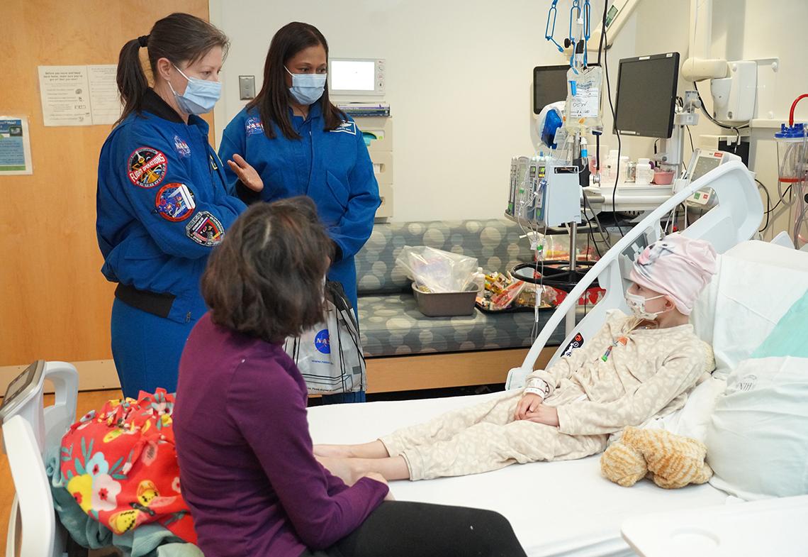 Two astronauts stand by pediatric patient lying in bed.