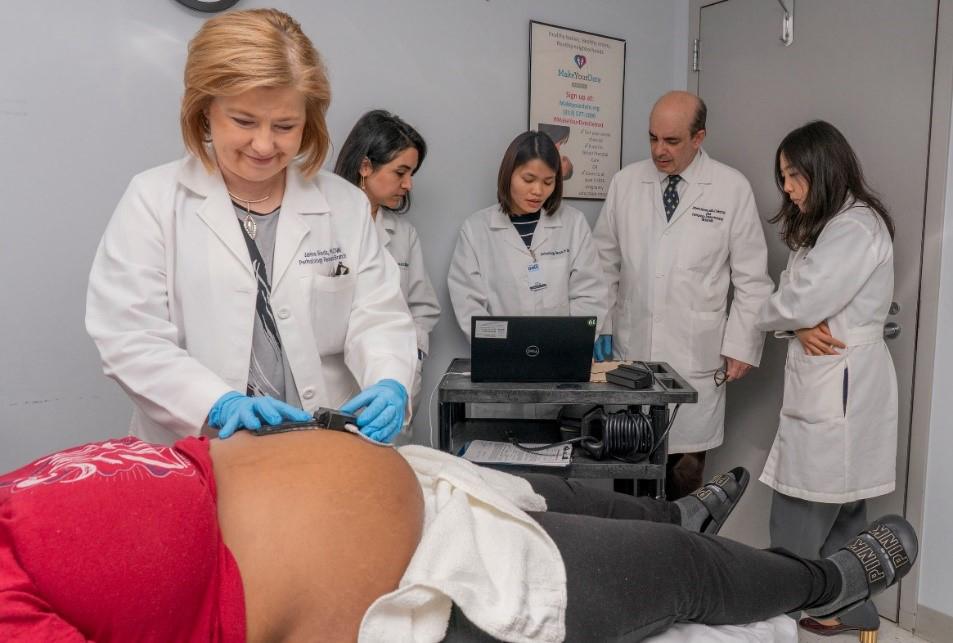 A smiling doctor holds the device against a pregnant patient's stomach with gloved hands. In the background, four other individuals in white coats gather around a laptop where the biosensor relays the data it collected.
