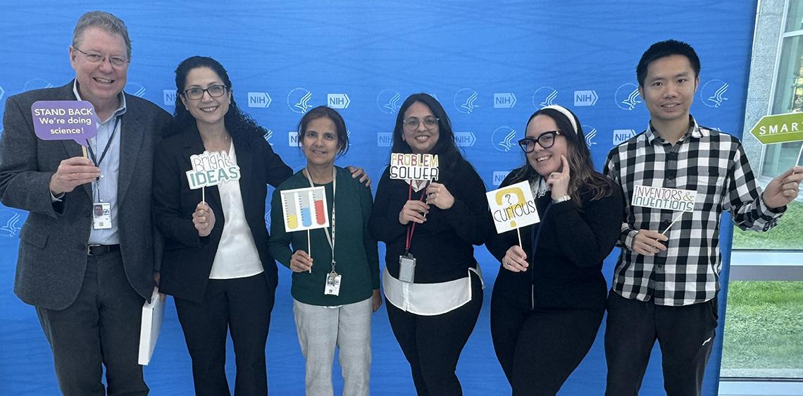 Six smiling colleagues pose in front of NIH backdrop holding such signs as: "Bright Ideas" and "Stand back: we're doing science."