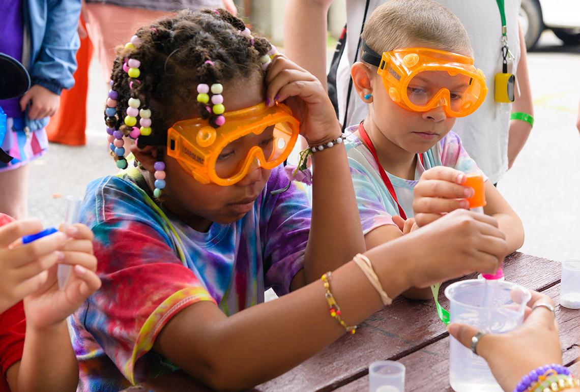 Two kids in goggles mix a solution in a cup.