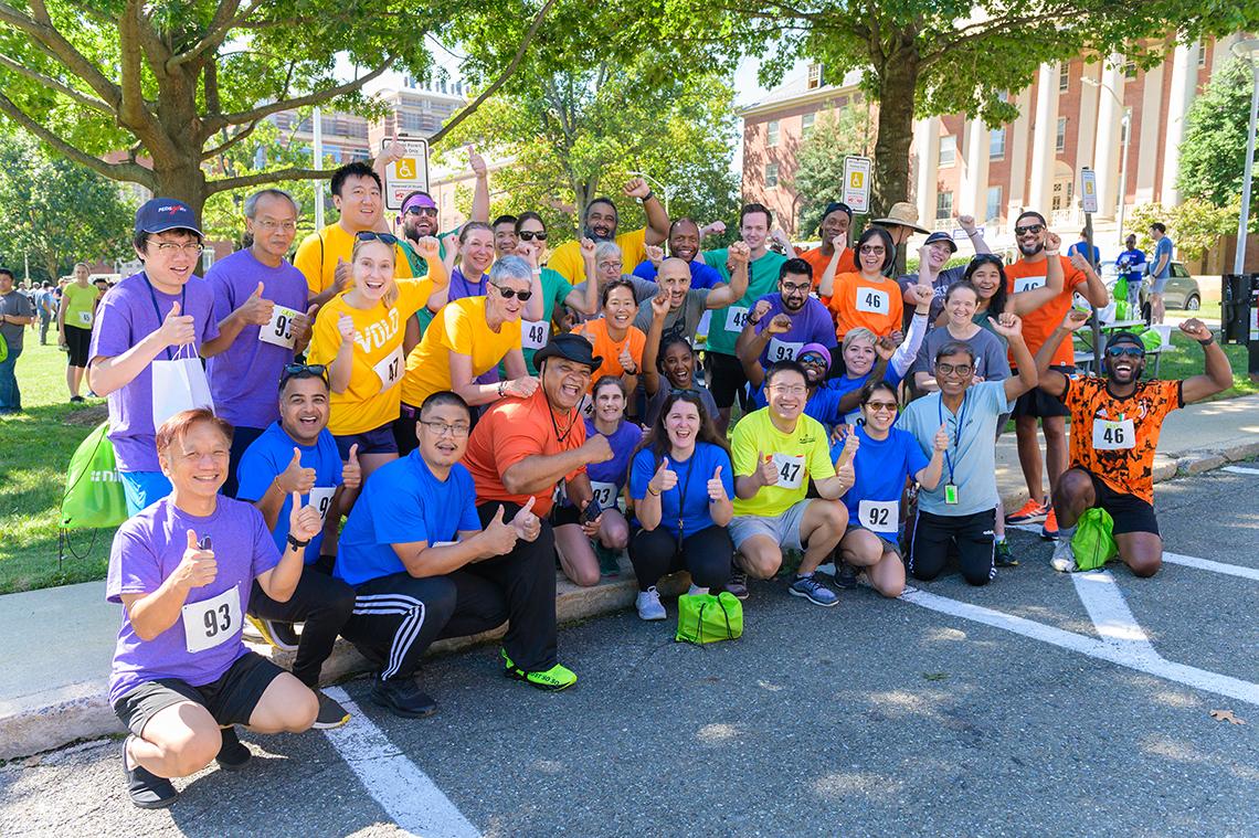 Several teams, each in colorful race-day shirts, pose together outside Building 1.