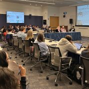Attendants at a horseshoe-shaped table, with more rows of participants in chairs around the perimeter of the room. A projector screen is visible in the corner of the frame.